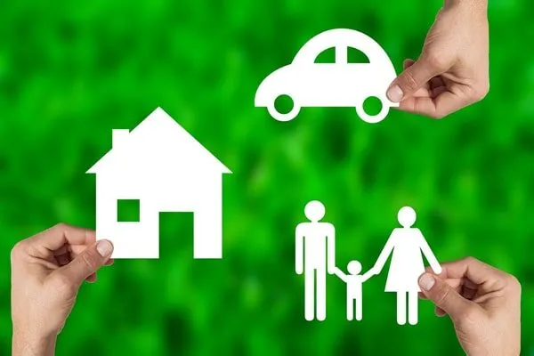 Mani che reggono sagome di carta a forma di casa, auto e famiglia su sfondo verde, simbolo di polizze assicurative